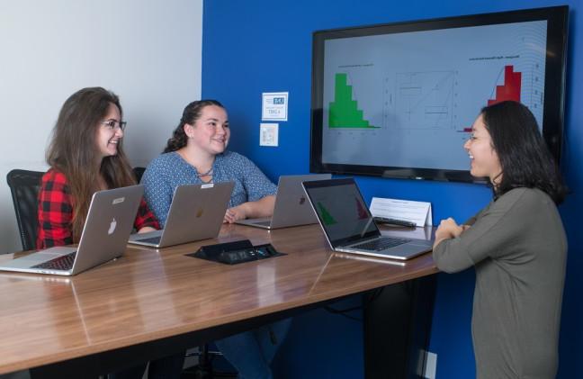 一群数据科学专业的学生坐在一张桌子旁，桌上放着他们的笔记本电脑和大显示器上的图表